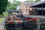 Several narrowboats are moored along a canal, bordered by a brick pathway and surrounded by lush greenery. A historic brick building and a metal bridge are visible in the background, adding an industrial yet tranquil atmosphere.