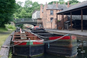 Several narrowboats are moored along a canal, bordered by a brick pathway and surrounded by lush greenery. A historic brick building and a metal bridge are visible in the background, adding an industrial yet tranquil atmosphere.