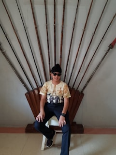 A person is sitting on a throne-like chair made of wood and spears arranged in a fan shape behind them. They are wearing sunglasses, a black cap, a printed t-shirt, dark pants, and sneakers.