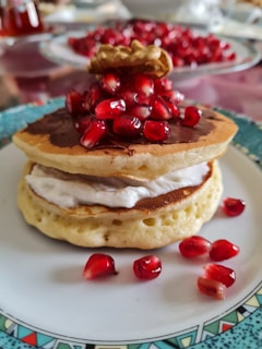 A stack of pancakes is topped with a generous portion of pomegranate seeds and a walnut. The pancakes are served on a decorative plate with a pattern around the edge. The stack has layers of cream and chocolate spread in between, and additional pomegranate seeds are scattered on the plate.