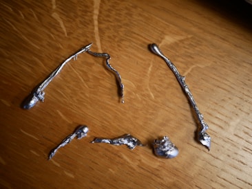 Several irregularly shaped metallic pieces are scattered on a wooden surface. The objects have a shiny, silver appearance and varied forms with twisted and elongated shapes.