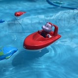 A small plastic toy boat is floating in clear blue water. The boat is red and features a cute grey animal figure, resembling a hippopotamus, seated inside. Surrounding the boat are other colorful toy sea creatures, including a blue fish with green details and a small orange creature.