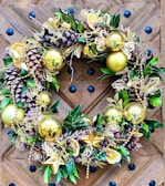 A decorative wreath adorned with golden ornaments, pine cones, and lush green leaves. The design includes gold baubles, star shapes, and dried citrus slices, complemented by golden beads draped throughout. The wreath is placed on a wooden door featuring a chevron pattern with black decorative studs.