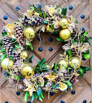 A decorative wreath adorned with golden ornaments, pine cones, and lush green leaves. The design includes gold baubles, star shapes, and dried citrus slices, complemented by golden beads draped throughout. The wreath is placed on a wooden door featuring a chevron pattern with black decorative studs.