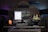 A modern home office setup featuring a clean white desk. A large monitor in the center displays a split-screen of text documents and code, with a laptop to the right showing an image of a vintage car. The desk is illuminated by two floor lamps on either side, casting a purple and blue ambient light. There are decorative items including a small cactus, a Himalayan salt lamp, and a green mug. Above the desk are three framed photographs: one of a vintage car, another of a beach, and the third is abstract art.