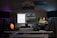 A modern home office setup featuring a clean white desk. A large monitor in the center displays a split-screen of text documents and code, with a laptop to the right showing an image of a vintage car. The desk is illuminated by two floor lamps on either side, casting a purple and blue ambient light. There are decorative items including a small cactus, a Himalayan salt lamp, and a green mug. Above the desk are three framed photographs: one of a vintage car, another of a beach, and the third is abstract art.