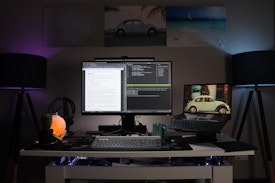 A modern home office setup featuring a clean white desk. A large monitor in the center displays a split-screen of text documents and code, with a laptop to the right showing an image of a vintage car. The desk is illuminated by two floor lamps on either side, casting a purple and blue ambient light. There are decorative items including a small cactus, a Himalayan salt lamp, and a green mug. Above the desk are three framed photographs: one of a vintage car, another of a beach, and the third is abstract art.