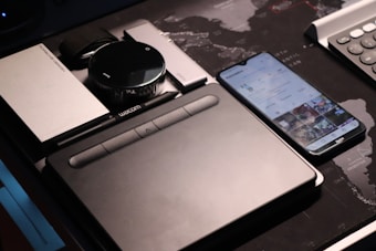 A collection of electronic gadgets is neatly arranged on a desk, including a drawing tablet, a smartphone displaying a social media app, a portable hard drive, and a camera lens. The surface underneath features a world map design.