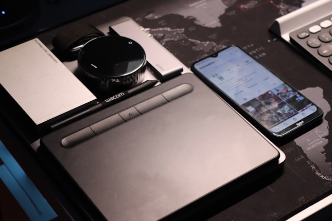 A collection of electronic gadgets is neatly arranged on a desk, including a drawing tablet, a smartphone displaying a social media app, a portable hard drive, and a camera lens. The surface underneath features a world map design.