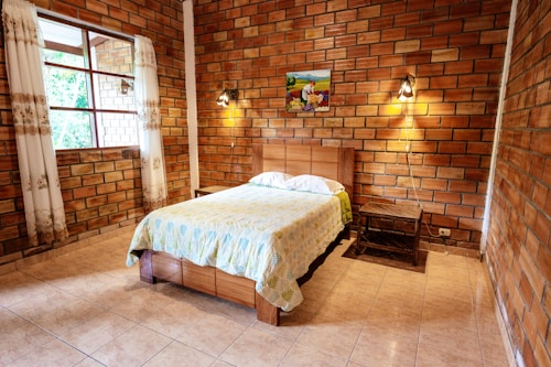 A cozy bedroom with brick walls, a wooden bedframe with a floral-patterned bedspread, and a small side table. A colorful painting is hung above the bed, with two wall-mounted lamps providing soft lighting. The large window, adorned with decorative curtains, lets in natural light, illuminating the warm-toned brickwork and tiled floor.