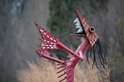 A whimsical metal sculpture resembling a dragon or fantastical creature, with a red color and white polka dots along its body. It features large, pointed teeth and black wire-like whiskers projecting from its face.