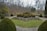 A well-maintained garden featuring circular trimmed hedges arranged on a raised stone planter. The garden is surrounded by a paved pathway bordered by symmetrical shrubs. In the background, a dense cluster of leafless trees creates a natural barrier. A green bench can be seen on the left side.