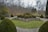 A well-maintained garden featuring circular trimmed hedges arranged on a raised stone planter. The garden is surrounded by a paved pathway bordered by symmetrical shrubs. In the background, a dense cluster of leafless trees creates a natural barrier. A green bench can be seen on the left side.