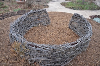 An outdoor setting features a circular structure made of intertwined branches. The structure resembles a large nest and is placed on a bed of mulch, surrounded by a garden path and some sparse vegetation.