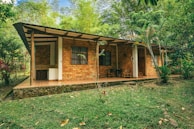 A rustic brick house with a sloping roof sits surrounded by lush greenery. The house features a shaded porch with a couple of chairs and a small table. Tall trees and abundant vegetation encircle the dwelling, giving it a cozy and secluded feel. The lawn in front is well-maintained, with some scattered leaves adding to the natural ambiance.