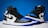 A pair of high-top athletic sneakers with a predominantly black, blue, and white color scheme is placed on a contrasting background. The shoes feature black laces and a distinctive logo on the side.