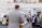 A man stands at a podium addressing a group of people in a spacious room with two levels. The audience, some wearing masks, is seated and standing, listening attentively. The setting suggests a formal gathering, possibly a community meeting or religious service.