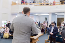 A man stands at a podium addressing a group of people in a spacious room with two levels. The audience, some wearing masks, is seated and standing, listening attentively. The setting suggests a formal gathering, possibly a community meeting or religious service.
