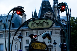 An ornate Art Nouveau style entrance to a subway station, featuring signs with the word 'Metropolitain' and 'Saint-Michel'. The background includes a historic building with a slate roof and intricate stone architecture. Lamp posts with decorative red lights add to the scene.