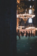 A spacious interior with intricate chandeliers hanging from the ceiling, featuring a large wall ornament with Arabic calligraphy. People are gathered in small groups on an ornate carpet, surrounded by grand architectural arches and lit by natural light streaming through large windows.