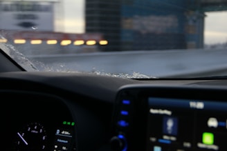 The image captures an interior view from a car, focusing on the dashboard and a partly frosted windshield. The dashboard displays a digital screen with various icons and gauges, while outside, blurred lights and structures create a sense of motion.