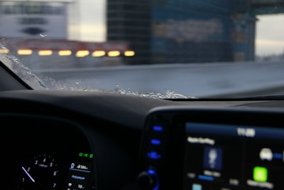 The image captures an interior view from a car, focusing on the dashboard and a partly frosted windshield. The dashboard displays a digital screen with various icons and gauges, while outside, blurred lights and structures create a sense of motion.