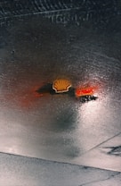 A glossy surface reflecting the yellow and red colors of a Shell gas station logo, creating an abstract, distorted appearance. The ground appears wet, possibly due to rain, enhancing the colors and reflective effect.