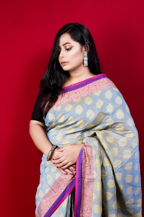 A woman with long black hair is wearing a traditional sari with a patterned design in shades of blue, gold, and pink. She is accessorized with dangling earrings and a bangle, standing against a vibrant red background. Her expression is calm and contemplative.