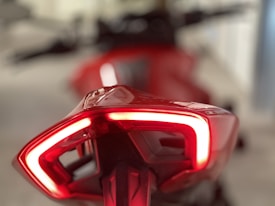 A close-up view of a sleek, red motorcycle tail light with a bright LED outlining its shape. The background is blurred, emphasizing the modern design and glossy finish of the tail light.