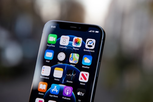 A smartphone displays a grid of app icons on its screen against a blurred background. The apps include FaceTime, Calendar, Photos, and others. The status bar shows a time of 9:41 and a 5G connection indicator at the top right.