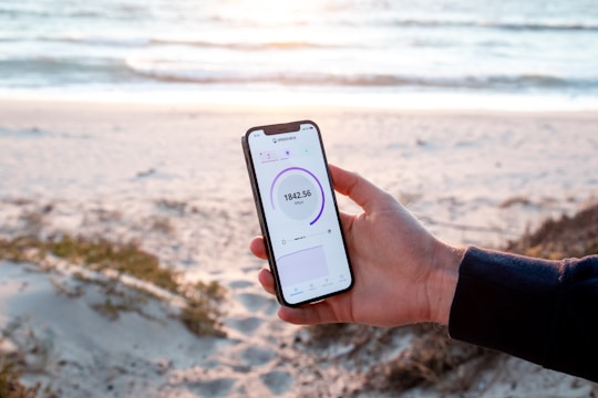 A person holds a smartphone displaying a speed test result while standing on a sandy beach. The ocean is in the background with gentle waves and a serene atmosphere. The phone's screen shows a speed of 184.56 Mbps and is held firmly by a hand wearing a dark sleeve.