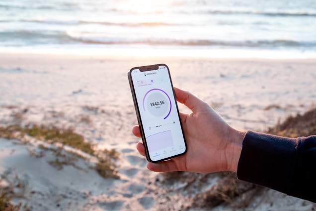 A person holds a smartphone displaying a speed test result while standing on a sandy beach. The ocean is in the background with gentle waves and a serene atmosphere. The phone's screen shows a speed of 184.56 Mbps and is held firmly by a hand wearing a dark sleeve.