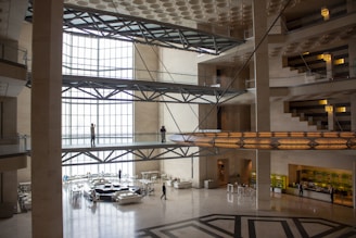 A spacious, modern interior with multiple stories connected by bridges. The architecture features large open spaces with glass walls allowing abundant natural light. Metal beams and geometric patterns are prominent in the design. Several people are visible, walking or standing on the bridges and the ground floor.