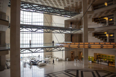 A spacious, modern interior with multiple stories connected by bridges. The architecture features large open spaces with glass walls allowing abundant natural light. Metal beams and geometric patterns are prominent in the design. Several people are visible, walking or standing on the bridges and the ground floor.