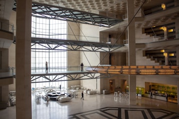 A spacious, modern interior with multiple stories connected by bridges. The architecture features large open spaces with glass walls allowing abundant natural light. Metal beams and geometric patterns are prominent in the design. Several people are visible, walking or standing on the bridges and the ground floor.