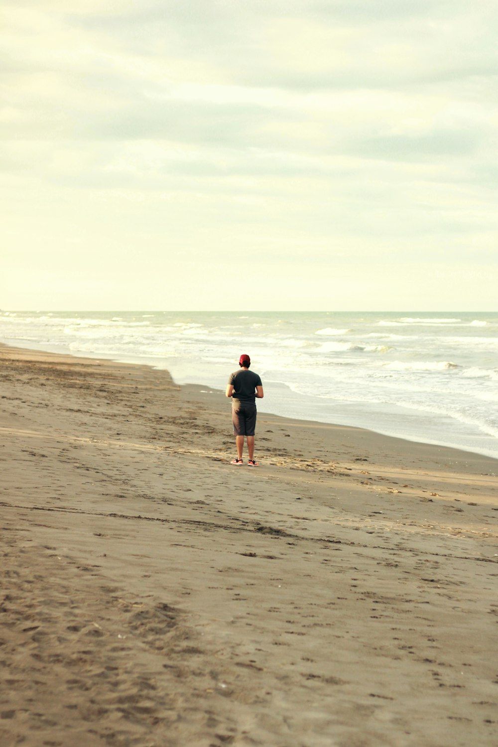 昼間、海近くの茶色の砂浜に立つ黒いジャケットと黒い短パンの男