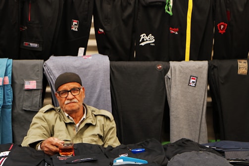 An elderly man wearing glasses and a cap sits in front of a display of folded pants from various brands including Reebok, Polo, and Fila. He is holding a glass with a dark liquid, likely tea. The pants are in different colors, such as black, grey, and blue, and prominently feature brand logos.