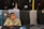 An elderly man wearing glasses and a cap sits in front of a display of folded pants from various brands including Reebok, Polo, and Fila. He is holding a glass with a dark liquid, likely tea. The pants are in different colors, such as black, grey, and blue, and prominently feature brand logos.