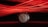 A wristwatch with a silver case and dark leather strap is centered in the foreground, featuring a white dial with Roman numerals and three subdials. Behind the watch, red light streaks create dynamic, parallel lines across a black background, casting a vivid glow.