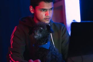 A young person is focused on a laptop screen while a small, curly-haired black dog with a blue bow tie sits nearby. The surroundings are dimly lit with a blue and red glow, creating a tech-savvy atmosphere.