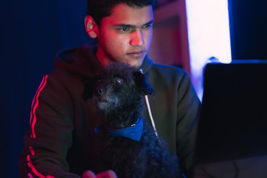A young person is focused on a laptop screen while a small, curly-haired black dog with a blue bow tie sits nearby. The surroundings are dimly lit with a blue and red glow, creating a tech-savvy atmosphere.