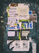 A weathered bulletin board covered with a variety of overlapping paper fliers and posters. The advertisements include rental listings, event notices, and service offers. The papers are in various stages of wear, with some crumpled and others torn. There is visible text in Spanish and a mix of graphics including logos and images. The background is a faded, chipped surface with graffiti around the edges.