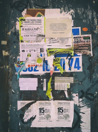 A weathered bulletin board covered with a variety of overlapping paper fliers and posters. The advertisements include rental listings, event notices, and service offers. The papers are in various stages of wear, with some crumpled and others torn. There is visible text in Spanish and a mix of graphics including logos and images. The background is a faded, chipped surface with graffiti around the edges.