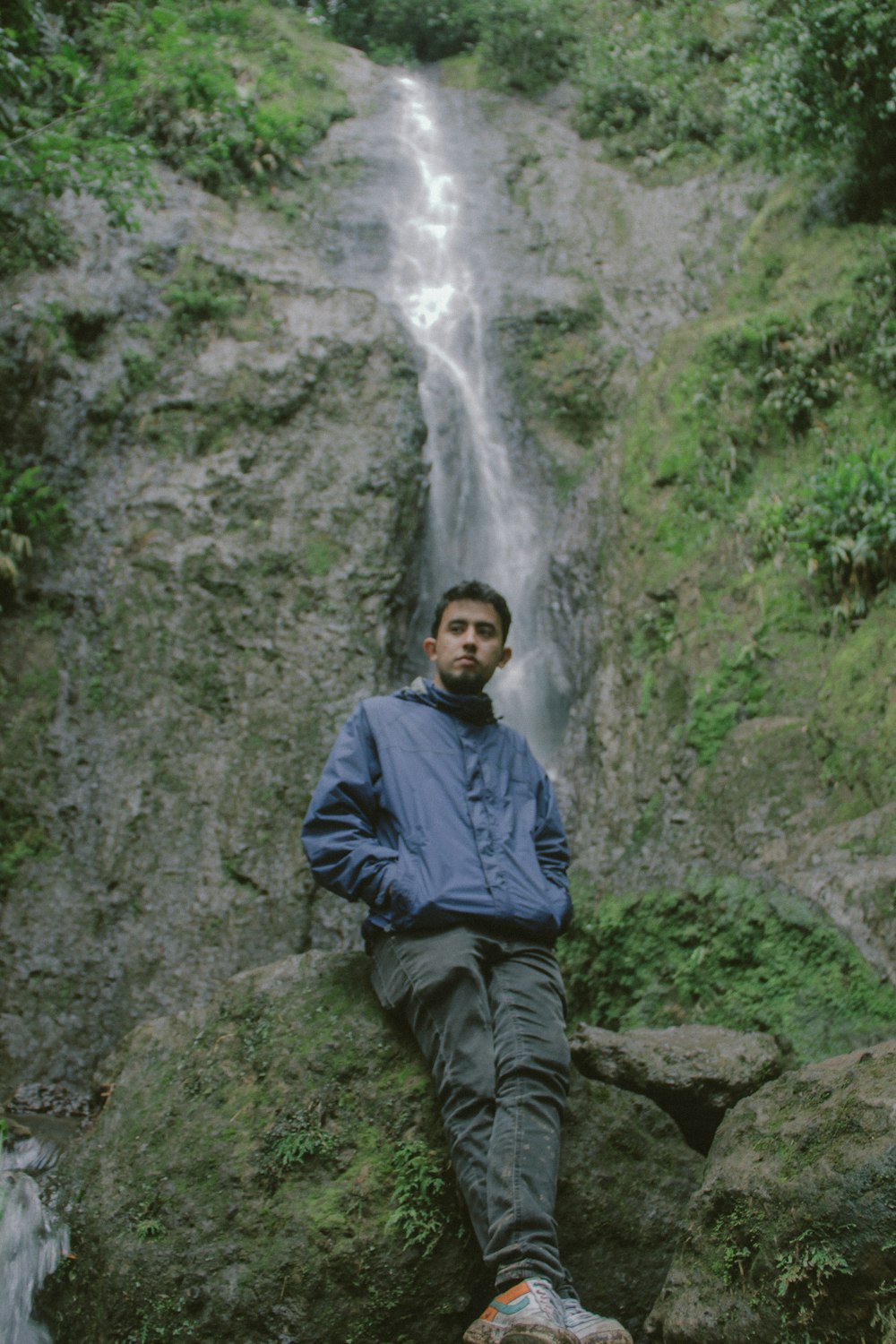 man in blue jacket sitting on rock near waterfalls during daytime