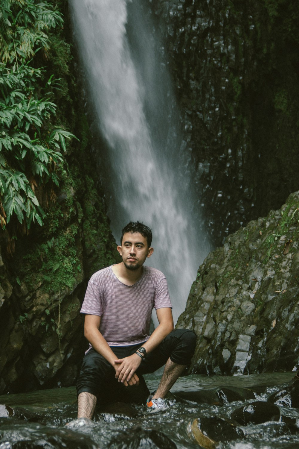 man in purple crew neck t-shirt sitting on rock near waterfalls during daytime