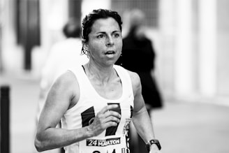 A person is running in a marathon or race, wearing a tank top with a visible race bib number. The background is blurred, indicating motion and activity.