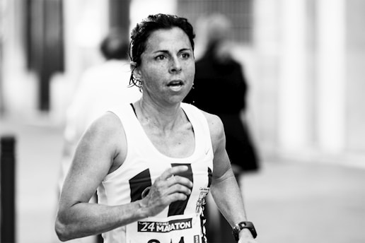 A person is running in a marathon or race, wearing a tank top with a visible race bib number. The background is blurred, indicating motion and activity.