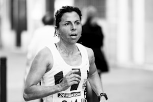 A person is running in a marathon or race, wearing a tank top with a visible race bib number. The background is blurred, indicating motion and activity.