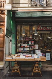 A quaint bookstore facade with a green awning, featuring a wooden table outside displaying a variety of books. The glass window behind showcases more books and has a warm interior light illuminating the shop.