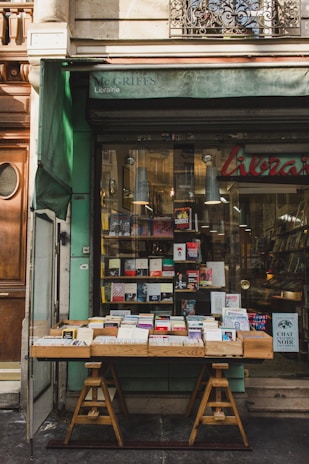A quaint bookstore facade with a green awning, featuring a wooden table outside displaying a variety of books. The glass window behind showcases more books and has a warm interior light illuminating the shop.